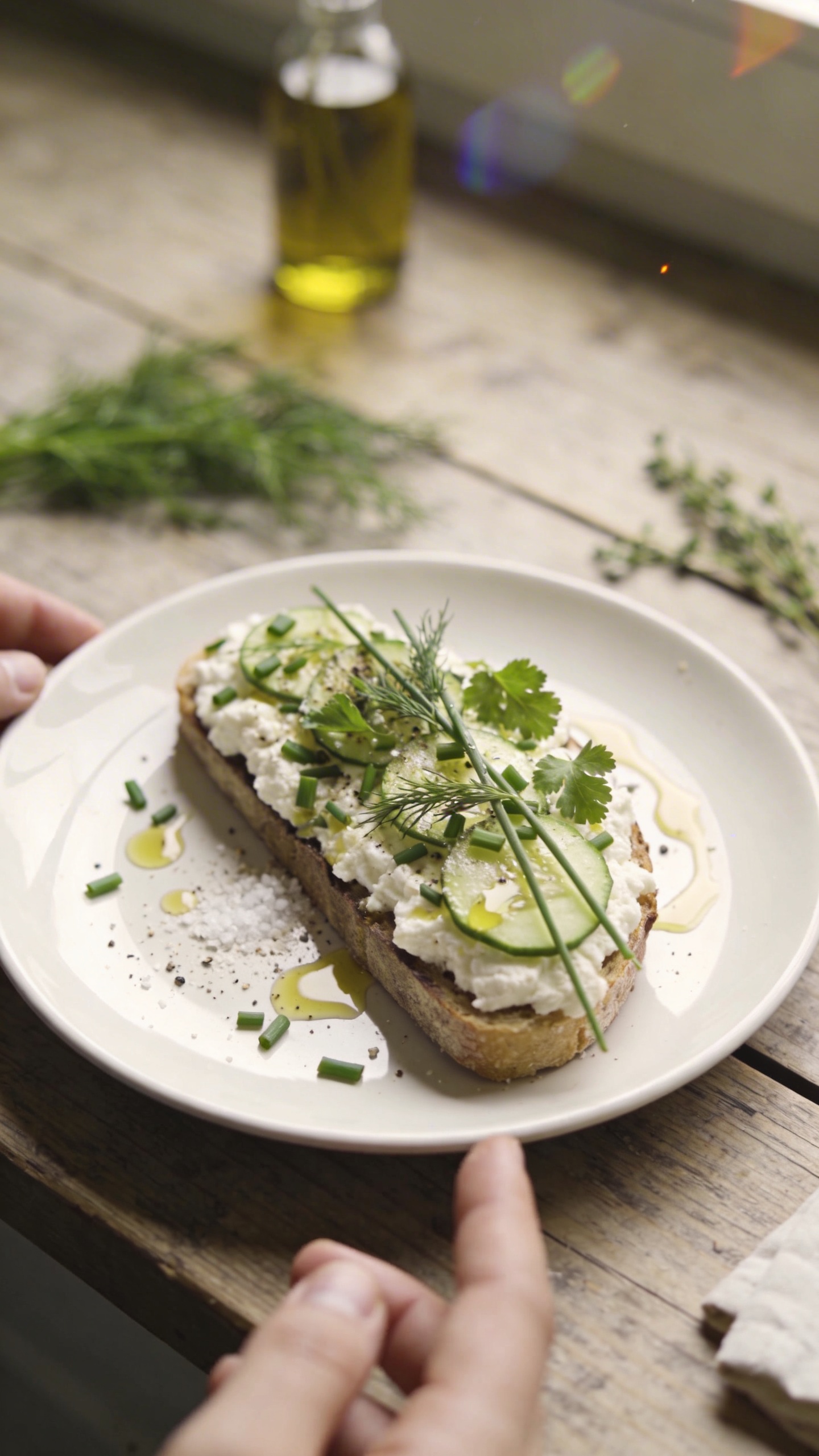 A real, authentic photo of a savory herb cottage cheese toast on a simple white plate resting on a rustic wooden kitchen table, captured as if by an iPhone in natural window light. The scene shows the toast topped with chopped chives, dill, and parsley, thin cucumber slices, a light olive oil drizzle, cracked pepper, and a pinch of sea salt, with a few herb sprigs and a small drizzle bottle in the background for context. The lighting is soft and natural from a nearby window, creating gentle shadows and a slightly overexposed highlight on the olive oil glaze. The composition is casual and slightly imperfect: a finger or two visible in frame, a faint reflection on the plate, a tilted horizon, and the edge of the table slightly out of alignment. Subtle motion blur on a napkin in the background, tiny grain visible in the lighter areas, and minor lens flare near the top corner to imitate real iPhone capture. The image should feel like a quick snack shot, no staging, no text, no professional lighting, with a compressed, 1080p quality look and a hint of post-processing that keeps it believable: slight smart blur with sharpening and a flat, natural color balance.