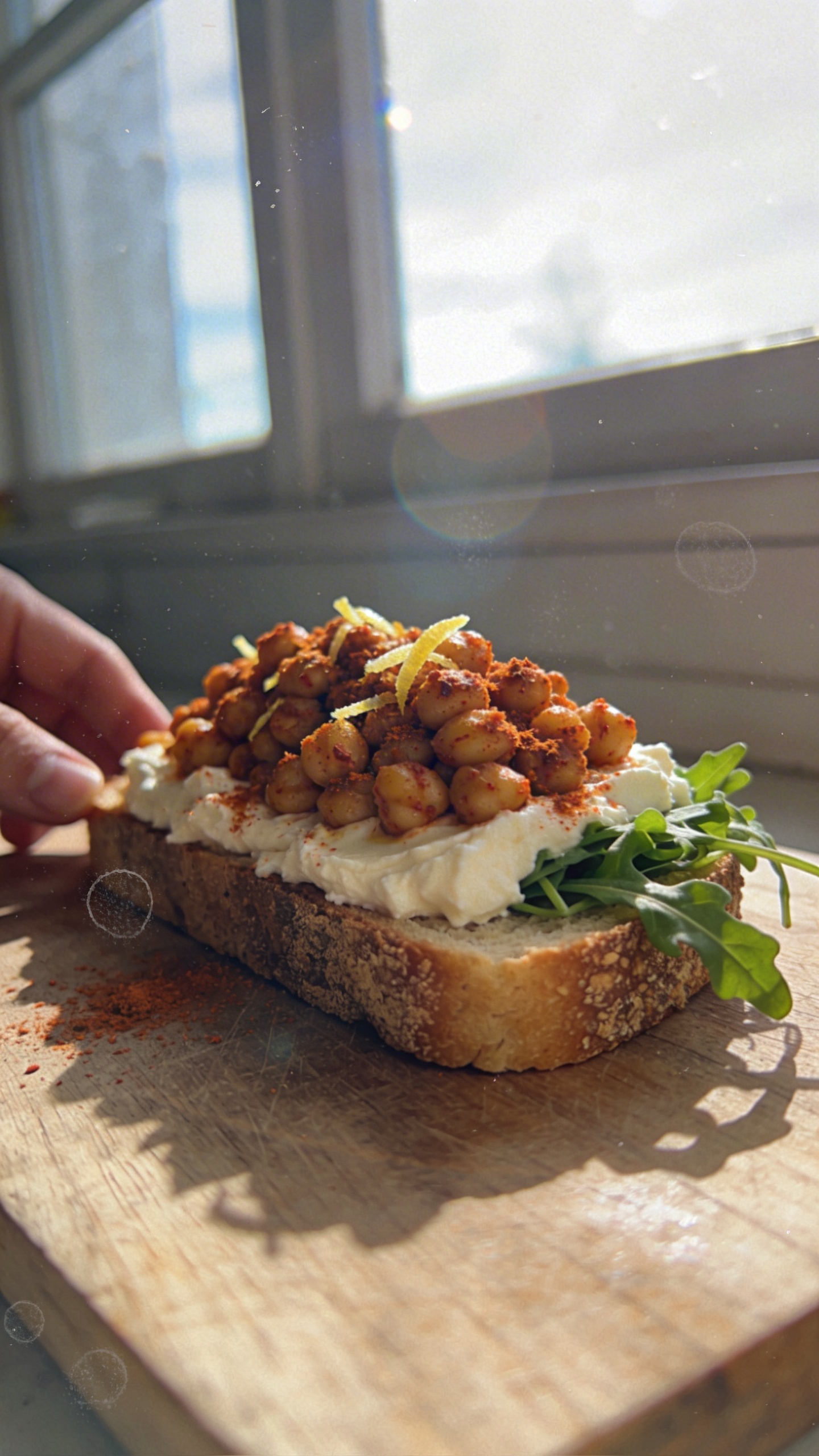 A photo-style prompt for an iPhone-shot image of a rustic toaster crusted with Smoky Chickpea Crunch on top of a lightly toasted slice, featuring roasted paprika chickpeas, a sprinkle of smoked paprika and lemon zest, a small handful of fresh arugula peeking from the edge, all laid on a casual wooden breakfast board. The main subject is the cottage cheese toast crowned with the smoky chickpea mixture, captured with natural window light streaming from one side to create soft shadows and a real-life, imperfect texture. Composition is casual and slightly uncentered, as if photographed quickly in a bustling kitchen or kitchen nook. Include subtle, realistic phone-camera imperfections: slight overexposed highlights from the window, a touch of lens flare near the edge, minor motion blur on a distant hand reaching for the toast, and grain that varies with the light. Horizon slightly tilted, some surface reflections on the plate, a fingerprint or smudge on the lens for authenticity. No staging, no obvious AI cues; the scene feels like a real moment: no words, no text overlays, “shot quickly” vibes, “taken in a hurry,” “fingerprint on lens,” “compressed, 1080p quality,” “screenshot quality.” Optional light post-processing hint: a subtle, offbeat adjustment that keeps it natural and not overly polished. The toast should clearly show the texture of roasted chickpeas with paprika, the pale creamiess of cottage cheese, and the bright green arugula, all in an authentic, down-to-earth photo.
