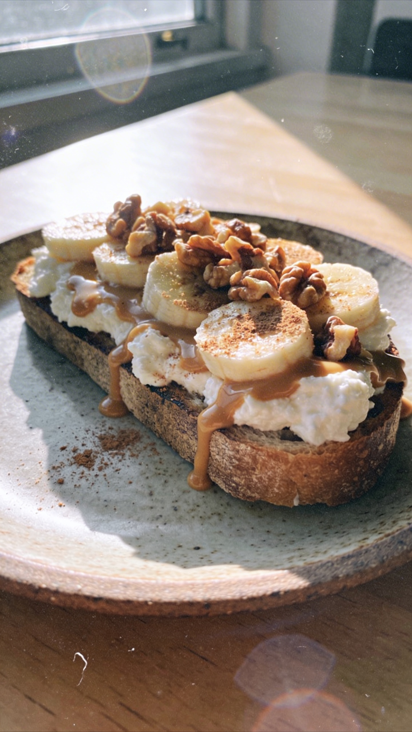 A candid, real-life photo of a Nutty Banana Split Toast as the main subject, placed on a rustic breakfast plate with a light wooden table in the background. The toast features a creamy cottage cheese spread topped with sliced bananas, crushed walnuts, and a drizzle of peanut butter, with a light cinnamon dusting for warmth. The scene should look like it was shot quickly in a sunlit kitchen window, with natural, soft window light, casual composition, and a slightly imperfect, realistic vibe. Include subtle, authentic phone-capture imperfections: slight overexposed highlights from the sun, gentle lens flare, tiny motion blur from a casual hand, and a touch of grain that varies with the ambient light. Horizon may be slightly tilted, and there should be minor fingerprints or smudges on the lens edge to emphasize realism. The image should resemble an ordinary iPhone photo, not staged or AI-generated, with compressed, 1080p quality and no staging cues. No text on the image. Optional subtle post-processing hint: a faint sense of reduced AI detection by light, natural edits (very slight contrast lift and softening) but still looking authentic and unpolished.