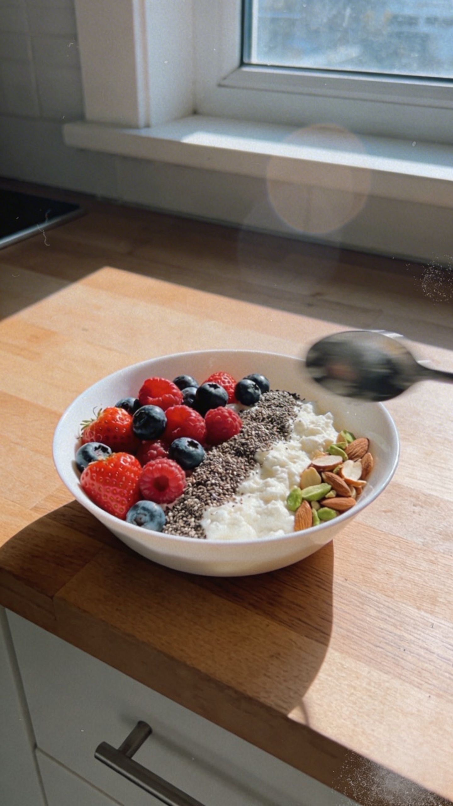 A real, authentic photo of a Cottage Cheese Energy Berry Bowl as the main subject, shot casually as if captured on an iPhone in natural window light. The bowl sits on a wooden kitchen counter near a sunny window, with soft, diffused daylight and gentle shadows. The bowl contains 1/2 cup cottage cheese topped with 1/2 cup mixed berries (strawberries, blueberries, raspberries) and a sprinkle of chia seeds, plus a few chopped almonds or pistachios on the side for crunch. Include a subtle, imperfect touch: slight overexposed highlights on the berries, a tiny lens flare from the window, a hint of motion blur on a spoon resting near the bowl, and a lightly tilted horizon of the counter. Graininess varies with light, and there may be a fingerprint or smudge on the lens in the corner. The composition is casual and unposed: no staging, no artificial props, and no text on the image. The shot feels like a quick, everyday moment—compressed, 1080p quality, no depth of field tricks or professional studio setup. Optional subtle post-processing hints: a tiny bit of processing that looks like “scaled down and up again” with light sharpening, and a flat color tweak that preserves natural textures.