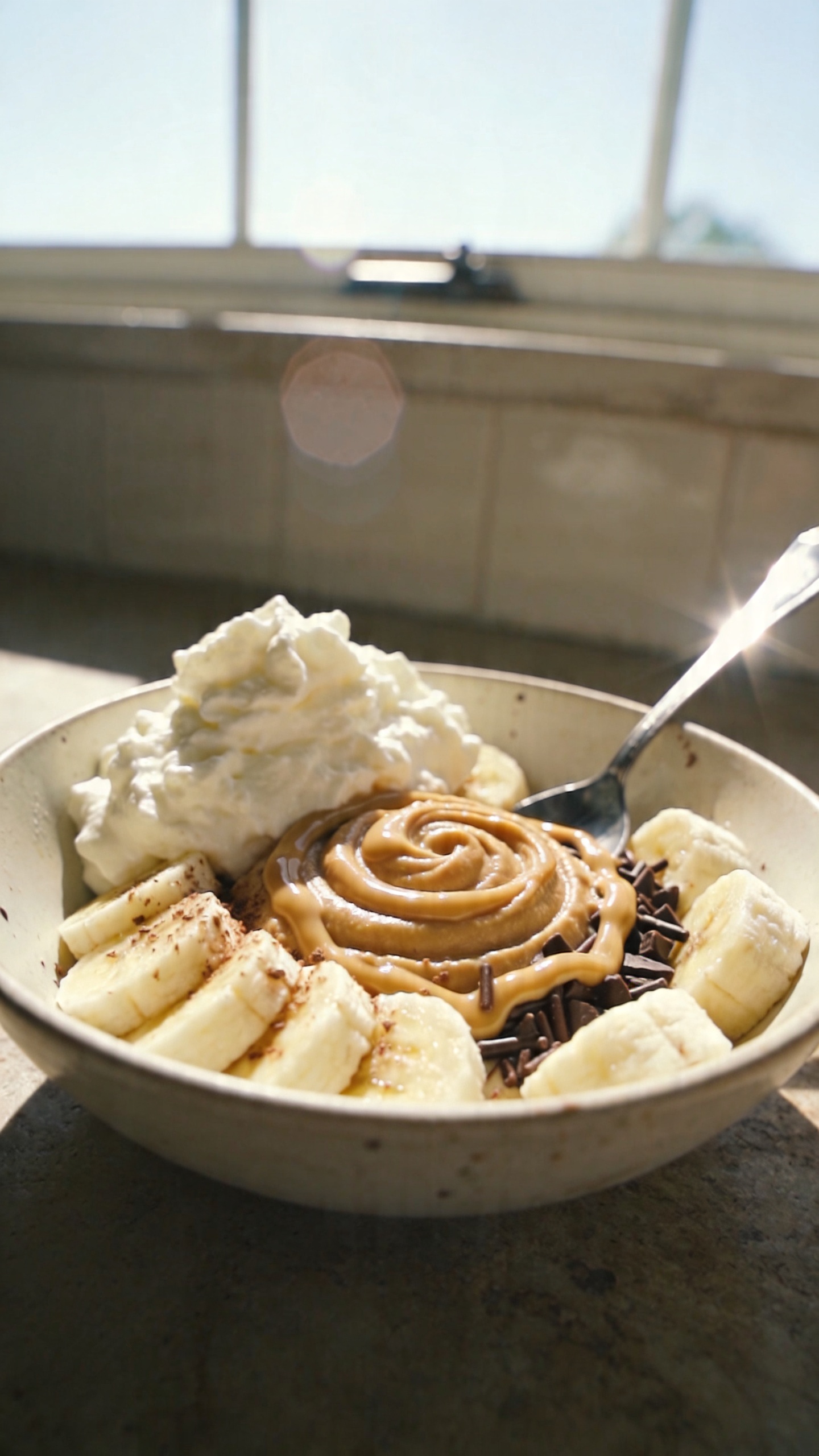 Take a natural, candid photo of a bowl of Banana Split Dream (Guilt-Free Edition) on a rustic kitchen counter, featuring creamy cottage cheese topping, frozen banana slices, and a triple-banana or almond butter swirl. The main subject is the bowl as the focal point, with visible elements: whipped cottage cheese topping, layered sliced bananas, and a drizzle or swirl of almond butter with a light sprinkle of dark chocolate. Include frozen banana slices around or tucked into the bowl for a vacation-in-a-bowl vibe. The scene should feel real and unposed, as if photographed quickly in a home kitchen with natural window light, capturing a casual composition and slight imperfections: a few overexposed highlights on the glossy spoon, a subtle fingerprint on the camera lens, a gentle tilt to the horizon, minor motion blur from setting the bowl down, and grain that varies with the light. The photo should look like iPhone-shot memory of a cozy breakfast moment—no staging, no text, no studio lighting, no dramatic depth of field. Keep it compressed 1080p quality, with a faint, natural lens flare from the window and a hint of post-processing that keeps it looking authentic rather than hyper-polished. No words on the image.