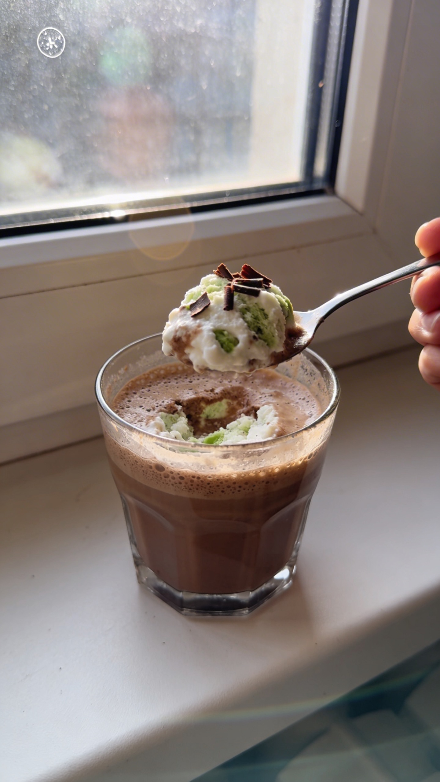A real, authentic photo of a spooned serving of Mocha Mint Snow in a Cup, featuring cottage cheese blended with espresso, cocoa, and mint note, presented in a clear glass cup with a frothy, mocha-tinted surface and a few chocolate shavings on top. The setting is casual and natural, taken with iPhone-style candid shot near a window with soft daylight. Include imperfect, realistic details: slight overexposed highlights from the window, subtle lens flare, a tiny fingerprint on the camera lens, minor motion blur from a casual hand pose, and a slightly tilted horizon. The composition should feel un-staged and spontaneous, with natural grain and a compressed, 1080p-like quality. No text on the image. Optional light post-processing hint: mild color tweaks that resemble a quick save or crop, but maintain a realistic, everyday look.