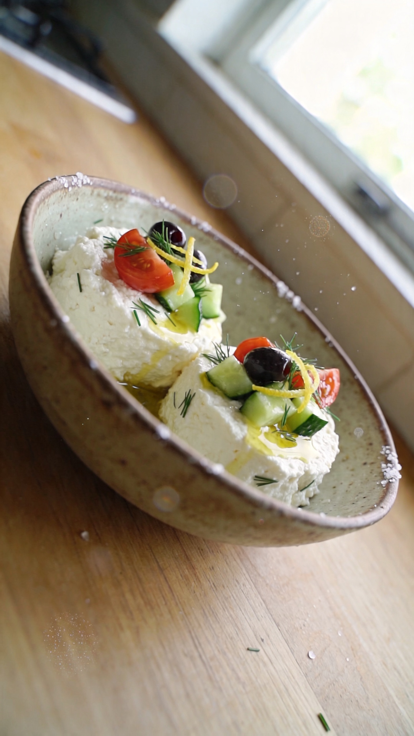 A photo of a savory cottage cheese bowl as the main subject, showcasing 2 cups of cottage cheese in a rustic bowl topped with chopped cucumber, cherry tomatoes, olives, and a dill or chive garnish, with a light olive oil drizzle and a hint of lemon zest. The scene is styled as a real, everyday meal: a casual, imperfect setup on a wooden kitchen table near a window, with natural daylight streaming in, slight reflections on the bowl, and a candid, unposed vibe. Include slight imperfections: a few grains of salt on the rim, minor condensation on the bowl, and subtle overexposed highlights from the window. Capture a moment as if photographed quickly with an iPhone—no staging, no studio lighting, and a slightly tilted horizon. Add realistic phone-camera quirks: tiny fingerprint marks on the lens, minor motion blur from a gentle hand shake, and grain that varies with light. Avoid any text in the image, and keep the look at “screenshot quality” feel with compressed, 1080p quality. Optional very light post-processing hints: a tiny, natural color punch without over-editing, reminiscent of an everyday, candid kitchen photo.