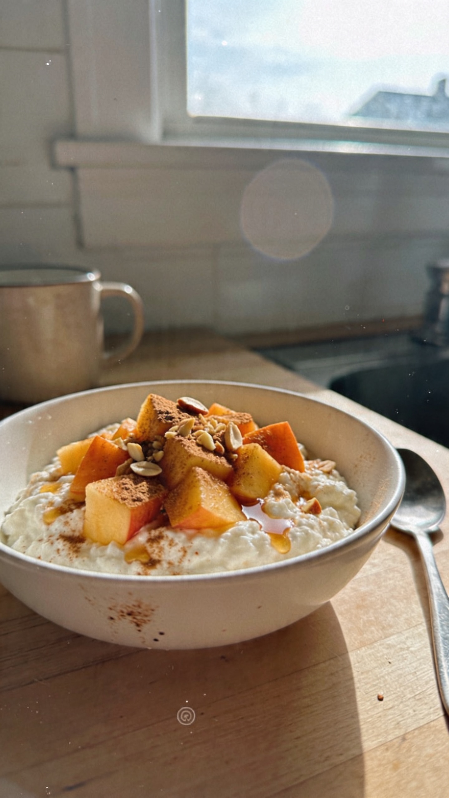 A candid, authentic-looking photo of a Creamy Cinnamon Peach Cottage Cheese Bowl as the main subject, placed on a casual breakfast setup. The bowl sits in a simple white ceramic dish on a wooden kitchen counter near a window, with natural morning light streaming in. The cottage cheese is creamy, topped with diced peaches, a light dusting of cinnamon, a splash of vanilla, and a few crushed almonds for crunch, visible texture and gloss from the natural light. The scene includes a casually placed spoon, a softly blurred mug in the background, and a lightly imperfect surface texture. The photo has a real, unposed vibe: slight overexposed highlights on the peach pieces, a tiny lens flare from the window, minor micro-scratches or smudges on the dish and counter, and a subtle tilt of the horizon. Slight graininess and compression to resemble 1080p phone-quality imagery. No text on the image. Shot quickly, no staging, fingerprint on the lens in the corner, no deep depth of field, as if taken with an iPhone in natural window light, with a natural, slightly imperfect, real-world feel. Optional very light post-processing that keeps it looking like a casual, real photo.