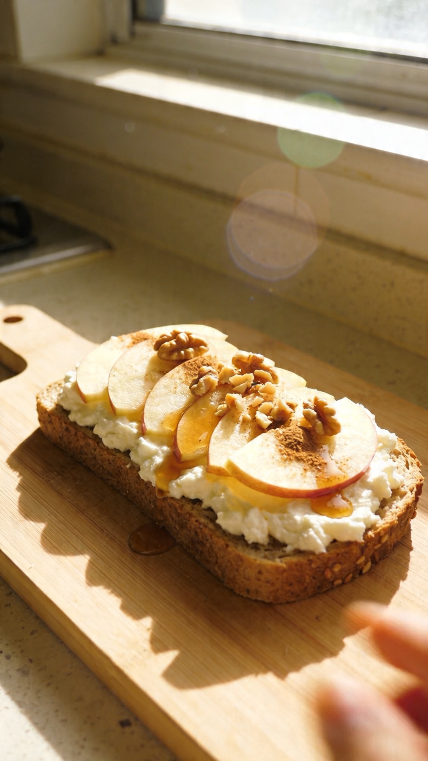 A real, authentic photo of Maple-Walnut Cinnamon Cottage Cheese Toast as the main subject, shot on an iPhone with natural window light. Casual, slightly imperfect composition: a wooden breakfast board on a kitchen countertop, maple-yellow light pooling from a nearby window, a slice of whole-grain toast spread thickly with cottage cheese, topped with thin apple slices, a light drizzle of maple, a pinch of cinnamon, and crushed walnuts for crunch. The scene should feel like a quick, no-staging morning setup: slight overexposed highlights on the toast, gentle lens flare from the window, a touch of motion blur from a quick pass of a hand, natural grain visible in the shadows, and a slightly tilted horizon. The image should look compressed, 1080p quality, with fingerprint smudge on the phone camera implied by the subtle blur, and a hint of post-processing to give a lived-in feel (scaled down and up again, slight smart blur with sharpening, recolored with a flat paint bucket). No text in the image.