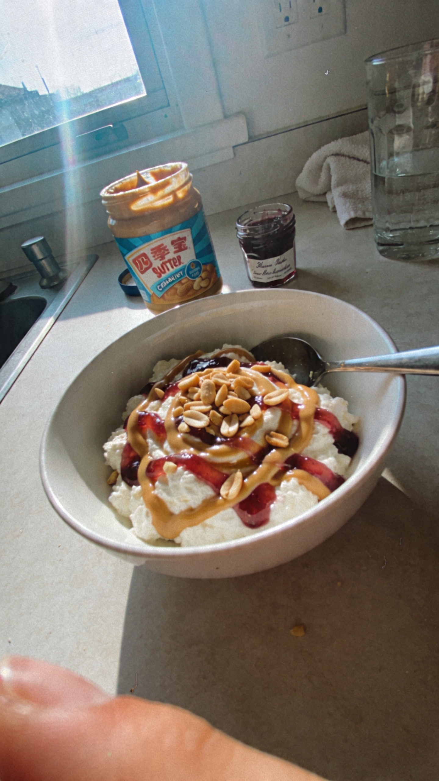 A candid, real-life photo of a bowl of Peanut Butter Jelly Mash-Up dessert inspired by cottage cheese-based recipes. The shot features a simple white ceramic bowl filled with creamy cottage cheese swirled with peanut butter and ribbons of fruity jelly, sprinkled with chopped peanuts for crunch. The main subject should be the bowl on a clutter-free kitchen counter near a window, with natural daylight streaming in to create soft, warm shadows. Include a few casual touches: a spoon resting in the bowl, a jar of peanut butter and a small jar of jelly in the background, and perhaps a partially visible glass of water or a towel. The image should feel authentic and un-staged, as if photographed quickly after preparing the snack. Style notes: - Shot quickly, no staging, casual composition - Slightly overexposed highlights from window light, natural lens flare - Minor motion blur or a finger on the edge of the frame for realism - Graininess that matches mid-mobility smartphone cameras, with a slightly tilted horizon - No text overlays or captions on the image - Compressed, 1080p quality feel, like a real iPhone capture - Optional subtle post-processing hints: muted color balance, gentle sharpening, but avoid obvious AI enhancements or perfect digital polish Main subject: the bowl of Peanut Butter Jelly Mash-Up dessert, clearly visible and centered enough to convey the dish, with the texture of cottage cheese, peanut butter swirls, jelly ribbons, and peanut crunch. Ensure the scene reflects a cozy, homemade post-workout or weekend snack vibe. Avoid any words in the image.