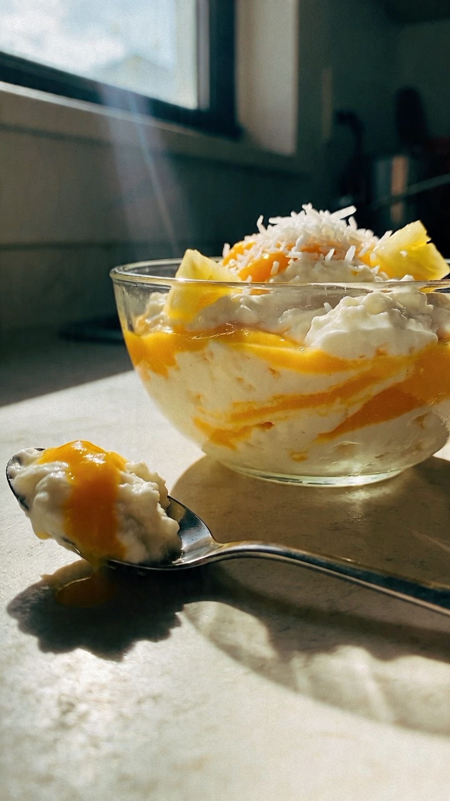 A real, authentic photo of a creamy tropical mango coconut cottage cheese dessert served in a simple glass bowl, with a visibly mango puree swirled into cottage cheese, and optional shredded coconut and a few pineapple bits for zing. The bowl sits on a casual kitchen counter near a window, with natural sunlight streaming in, creating soft, warm highlights and gentle shadows. Include a faint reflection on the glass, slight overexposure on the rim, and a touch of lens flare from the window. The scene should feel unstaged: a quick grab-and-go moment, perhaps with a spoon resting nearby and a casually arranged spoonful taken with a natural, imperfect tilt. Slight motion blur on the edge of a spoon in mid-scoop, grainier texture in the shadows, and a horizon that slightly tilts to convey realism. The overall look is compressed, 1080p quality, as if shot quickly on an iPhone, with no visible text or branding. Optional subtle post-processing hints: faint, natural color slightly desaturated and a tiny touch of sharpening on the bowl to keep details crisp without looking artificial. No words or captions in the scene.