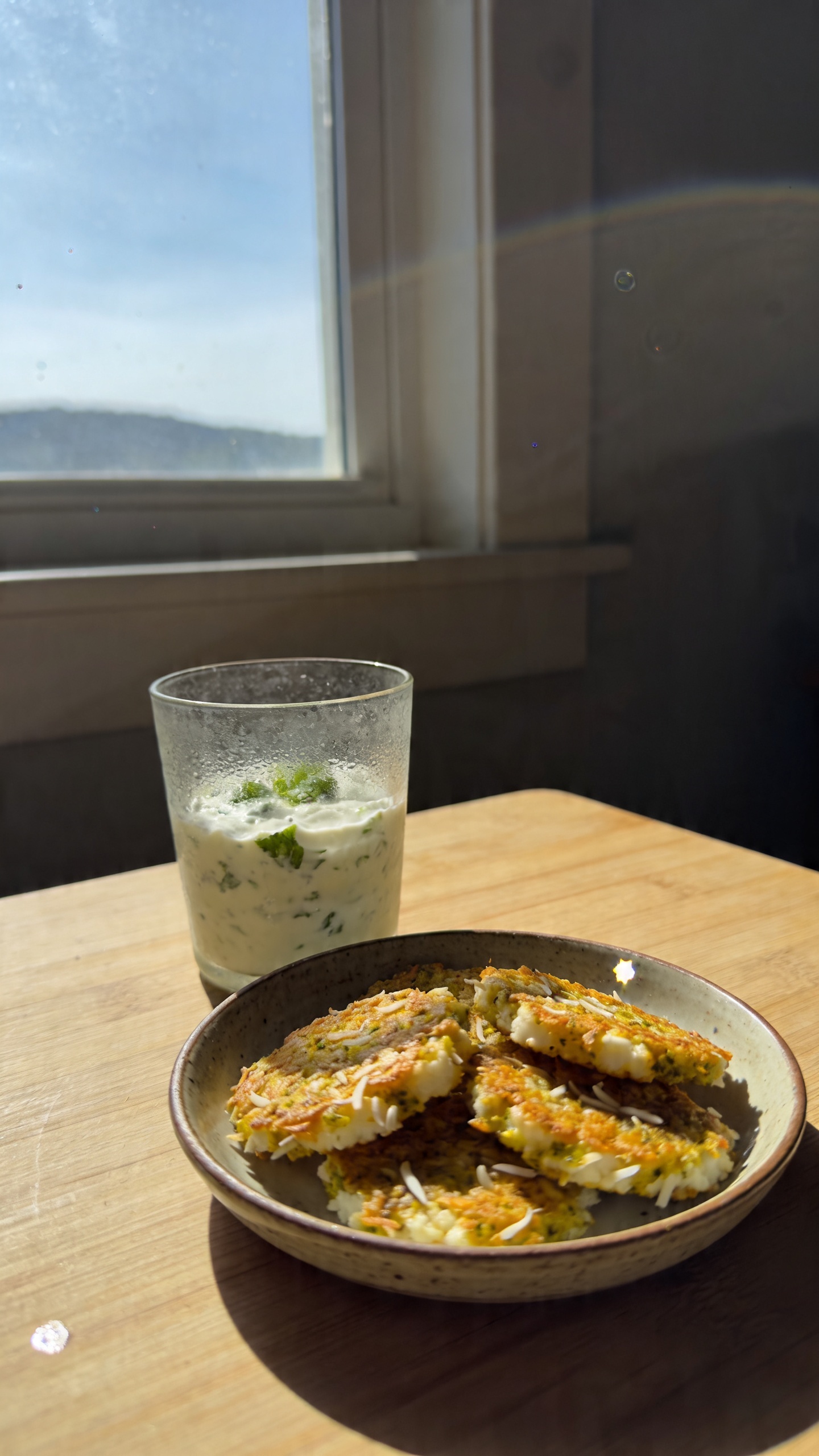 A real, authentic photo of coconut-curry cottage cheese crisps as the main subject, shot as if by an iPhone in natural window light. Casual, slightly imperfect composition: a small, rustic plate or bowl with crisp, golden coconut-curry crisps resting on a simple wooden table beside a lightly frosted glass of yogurt-mint dip for contrast. The scene feels un-staged and candid, as if grabbed between bites. Soft, ambient daylight from a nearby window creates gentle highlights and warm tones on the crisps, with natural shadows. Subtle, realistic imperfections: a tiny lens flare at the edge from the window, a hint of overexposed highlight on the rim of the dish, slight motion blur suggesting a quick grab, and a faint grain that varies with the light. The horizon is slightly tilted, adding to the casual feel. No text overlays or labels, no artificial studio setup. The image should look like compressed, 1080p quality, captured quickly, with no heavy post-processing—maybe a light, natural warmth but not polished. No words, only the food and setting, conveying a snack-prep vibe after a workout, with a hint of a tropical-curry aroma in the air. Avoid any cinematic or studio indicators; keep it as a real-life, everyday kitchen moment. Include a small fingerprint on the lens and a touch of color drift from natural light to enhance authenticity.
