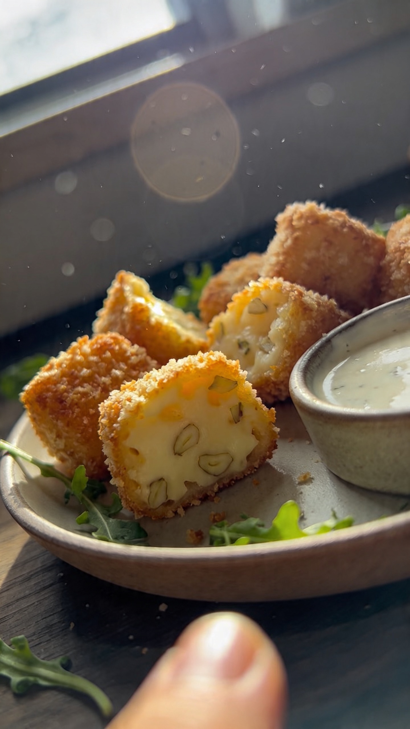 A close-up, realistically photographed bite-sized garlic parmesan snowflakes made with an air fryer, featuring a crisp, golden parmesan crust that visibly coats a tender interior with garlic flecks. The scene should center on a small plate or dipping dish of the cheesy bites, arranged casually among a few scattered green salad leaves or herbs for a natural, home-cooked vibe. The photo should look like it was taken with an iPhone, capturing a real, imperfect moment: natural window light streaming in, slight overexposed highlights on the cheese edges, soft lens flare from the sun, a touch of motion blur from a hurried snap, and a light grain or noise that increases in darker areas. The horizon should be subtly tilted, with a slightly off-kilter angle and a casual, unstyled background (kitchen counter or wooden table) to imply a spontaneous shot. No staging or studio setup; no visible branding or logos. The image should feel intimate and approachable, as if someone snapped it quickly while preparing a dinner starter. Include finger smudges or a fingerprint on the lens and minor, natural imperfections to enhance realism, with a compressed, 1080p quality look and no text overlays. Optional subtle post-processing hints: mild smart blur and sharpening in small areas, gentle color tweak to emphasize warm parmesan tones, but keep the overall natural, unpolished feel.