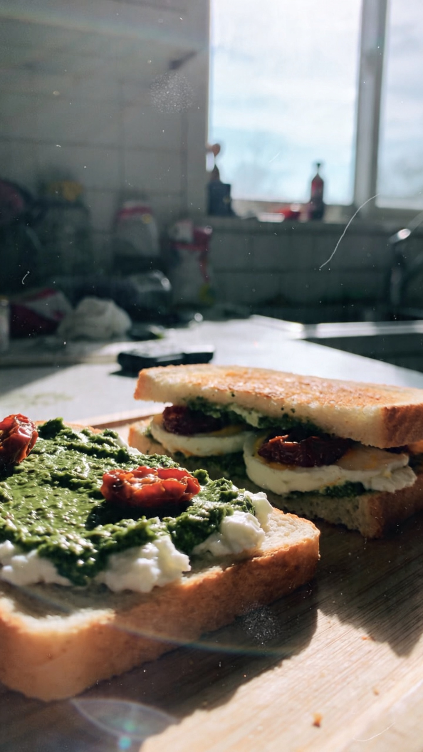 A real, authentic photo of an open-faced pesto cottage cheese breakfast sandwich on lightly toasted bread, showcased as the main subject. The scene is shot in natural window light with a casual, imperfect composition that feels quick and unstaged. Include a layer of bright green basil pesto on the bread, dollops of cottage cheese between slices, and sun-dried tomatoes for a tangy pop. The setting resembles a sunlit kitchen counter in the morning, with slight clutter and everyday items in the background to emphasize a lived-in feel. Capture subtle realism: slightly overexposed highlights from the window, gentle lens flare, a tiny bit of motion blur from a hand moving, and grain that varies with light. Horizon might be a touch tilted, and there could be fingerprint smudges on the lens. The image should read as “compressed, 1080p quality,” with no staging, no text, and no heavy post-processing that makes it look artificial. Optional light, imperfect post-processing hints: a slight smart blur with sharpening to mimic a quick edit, a muted color tweak that keeps natural tones, and a look that’s been scaled down and up again. No words, no branding, just a candid, sunlit pesto cottage cheese breakfast sandwich photographed as if taken with an iPhone in a real morning glance.