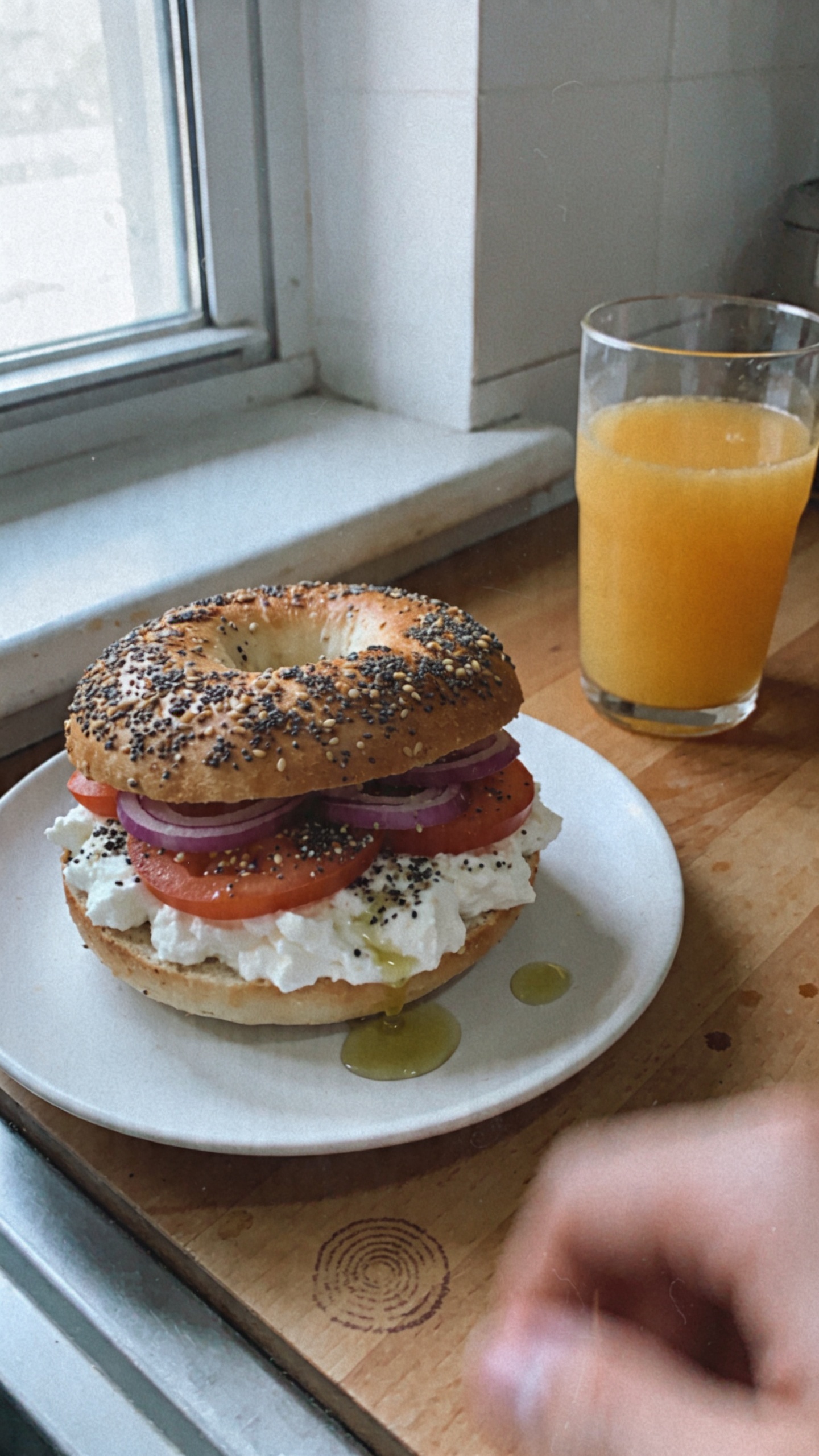 A casual, real-life kitchen breakfast scene featuring a toasted everything bagel spread with a generous layer of cottage cheese, topped with thin tomato slices and red onion, lightly sprinkled with everything bagel seasoning and a small drizzle of olive oil. The bagel sits on a simple white plate on a wooden kitchen counter next to a half-filled glass of orange juice. Natural window light pours in from the left, creating soft, imperfect shadows and gentle highlights on the cottage cheese. The shot is taken in a hurry, no staging, with a slightly tilted horizon to convey spontaneity. Subtle camera imperfections: a few overexposed highlights on the glass, slight fingerprint smear on the counter or lens, minor motion blur from a casual hand in the frame, and a bit of grain suggesting 1080p quality. The vibe is everyday, authentic, and unpolished, as if snapped quickly on an iPhone with no heavy editing—no text, no AI-esque polish, just a real breakfast moment. Optional light post-processing hints: slight color desaturation with a flat, phone-camera feel, a tiny bit of sharpening sparingly applied to edges, and a minimal warm tone to mimic natural kitchen lighting.