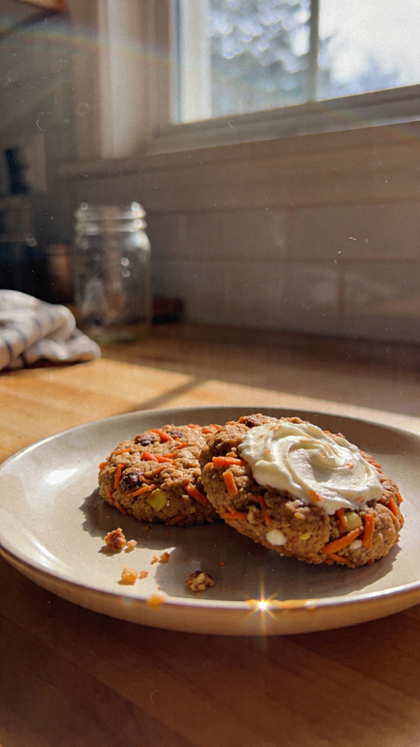 A real-looking photo of Carrot Cake Cookies with Cottage Cheese as the main subject, as if photographed for a casual food blog. The cookies should look moist and inviting, with visible shredded carrots and specks of warm cinnamon and ginger. Include a creamy swirl of cream cheese on one cookie for optional indulgence. Plate them on a simple, imperfect ceramic plate with a few crumbs around, set on a wooden kitchen table near a window. Use natural window lighting to create gentle shadows and a cozy, brunch-ready vibe. The shot should feel unposed and spontaneous: a quick, in-the-moment capture, not staged. Include subtle, camera-like imperfections: slight overexposed highlights on the plate edge, a touch of lens flare, minor motion blur in the background if any, and a faint grain that varies with light. Slightly tilted horizon, casual composition with one cookie stack or bite taken to imply real eating. Dim, real-world background with a hint of kitchen elements (mason jar, towel) but not cluttered. No text in the image. Shot quickly, taken in a hurry, fingerprint on lens, compressed, 1080p quality, screenshot-like quality, no cinematic or studio cues. Optional light post-processing hint: minimal, such as slight smart blur with sharpening and a flat color balance to avoid overly glossy look.