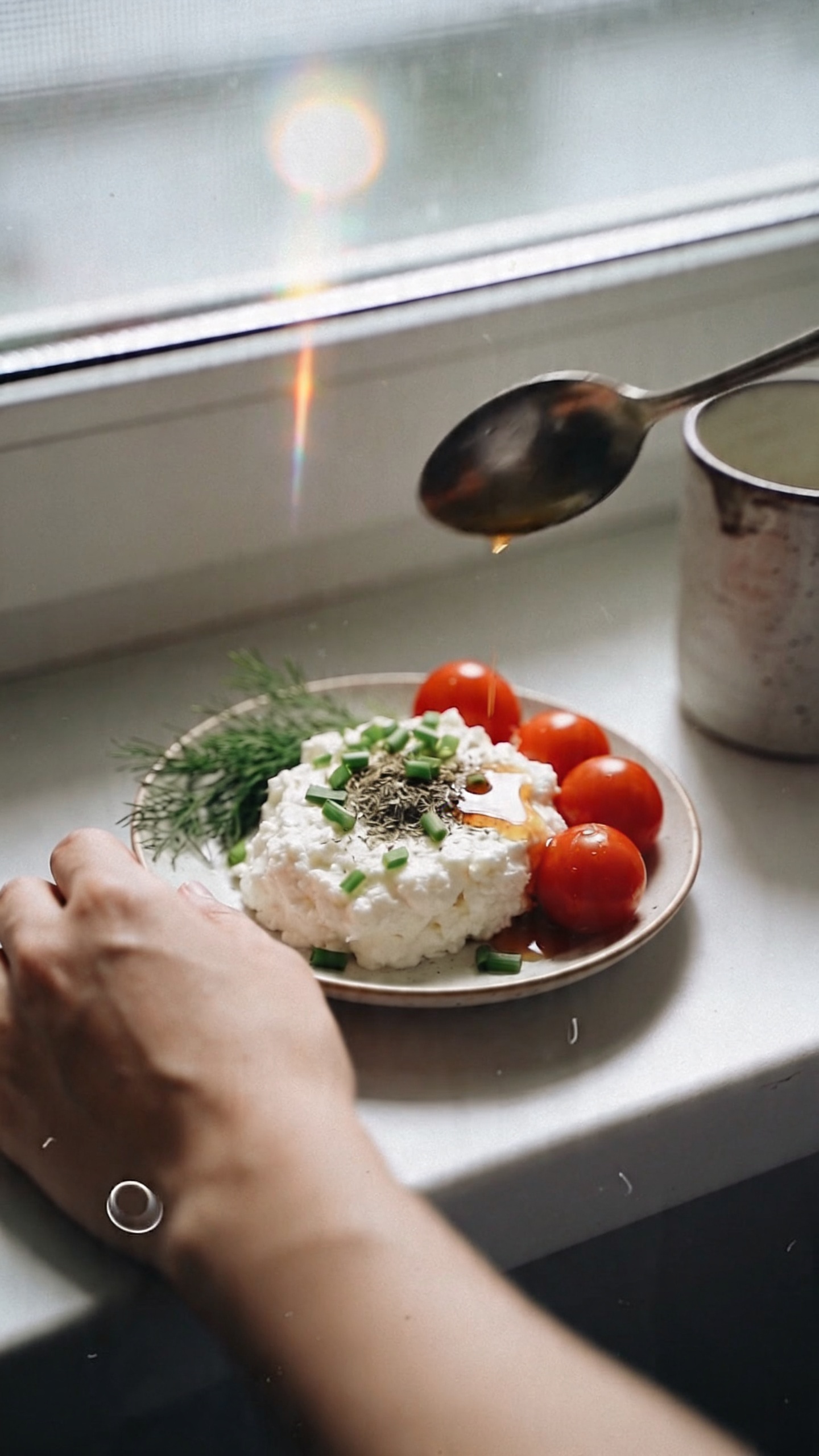 A casual, real-life kitchen breakfast scene featuring a person’s plate of Savory Herb Cottage Cheese Sunrise as the main subject, captured as if with an iPhone in natural window light. The bowl or small plate holds cottage cheese topped with chopped chives, a few dried dill or parsley sprigs, and bright cherry tomatoes, with a light splash of hot sauce or cracked pepper for color. The composition is shot quickly, in a no-staging, everyday-morning moment: a slightly imperfect, real-photo vibe with natural, soft window light, a candid hand resting nearby or a spoon in mid-air, and a casually placed mug or jar in the background. Include realistic phone-camera imperfections: slight overexposed highlights on the cottage cheese, a touch of lens flare from the window, minor motion blur from a quick, in-between-bite shot, and grain that varies with the lighting. The horizon line may be gently tilted, and there could be a fingerprint on the lens for authenticity. The scene should feel like a real, unglamorous morning capture, compressed to 1080p quality with no staged posing, no text, and no artificial studio setup. Optional subtle post-processing hints: slight smart blur with light sharpening and a flat color balance to resemble a casual, slightly processed phone photo.