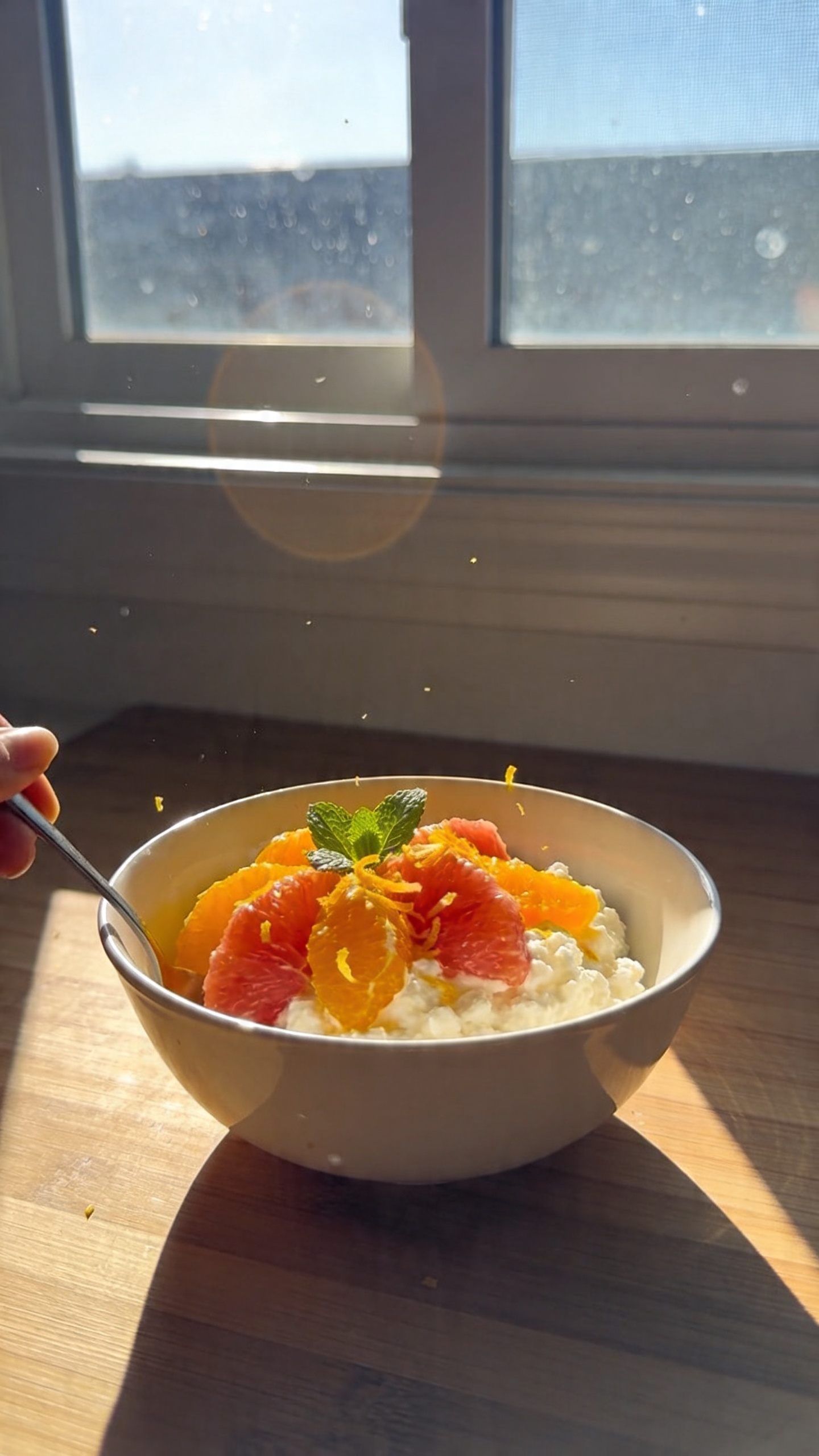A real, authentic photo of a Citrus Zest Sunrise Bowl for a weight-loss breakfast, shown in a casual, home-kitchen setting. The bowl sits on a wooden countertop near a window with natural daylight streaming in, creating soft, imperfect shadows. The cottage cheese base is bright and creamy, topped with vibrant orange and grapefruit segments, a light dusting of rind zest, and a few fresh mint leaves folded in. The composition is slightly off-center and un-staged, as if photographed quickly in a morning routine. Subtle camera imperfections are present: a hint of overexposed highlights on the citrus, a tiny lens flare from the window, a touch of motion blur from reaching for a spoon, and a faint grain that varies with the light. The horizon is slightly tilted, and there are tiny fingerprints or smudges on the lens, adding to the casual, realistic feel. The image should look like a compressed 1080p real photo, taken with an iPhone, with no text visible and no artificial studio setup. Optional subtle post-processing touches might include a mild, natural color lift and slight contrast adjustments to mimic quick, real-life editing, while preserving an authentic, no-staging vibe.